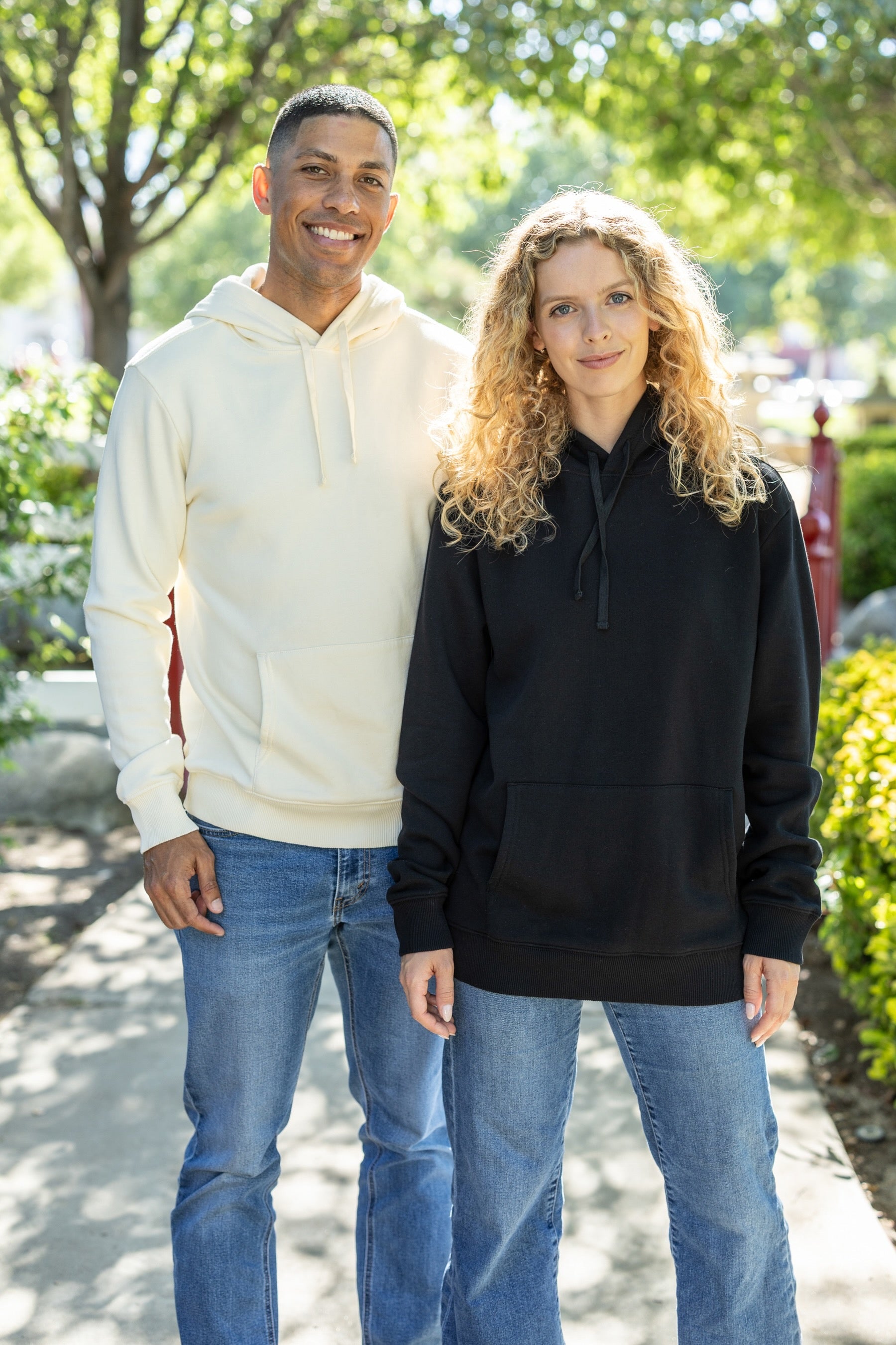 Two people wearing hoodies standing outdoors with greenery in the background