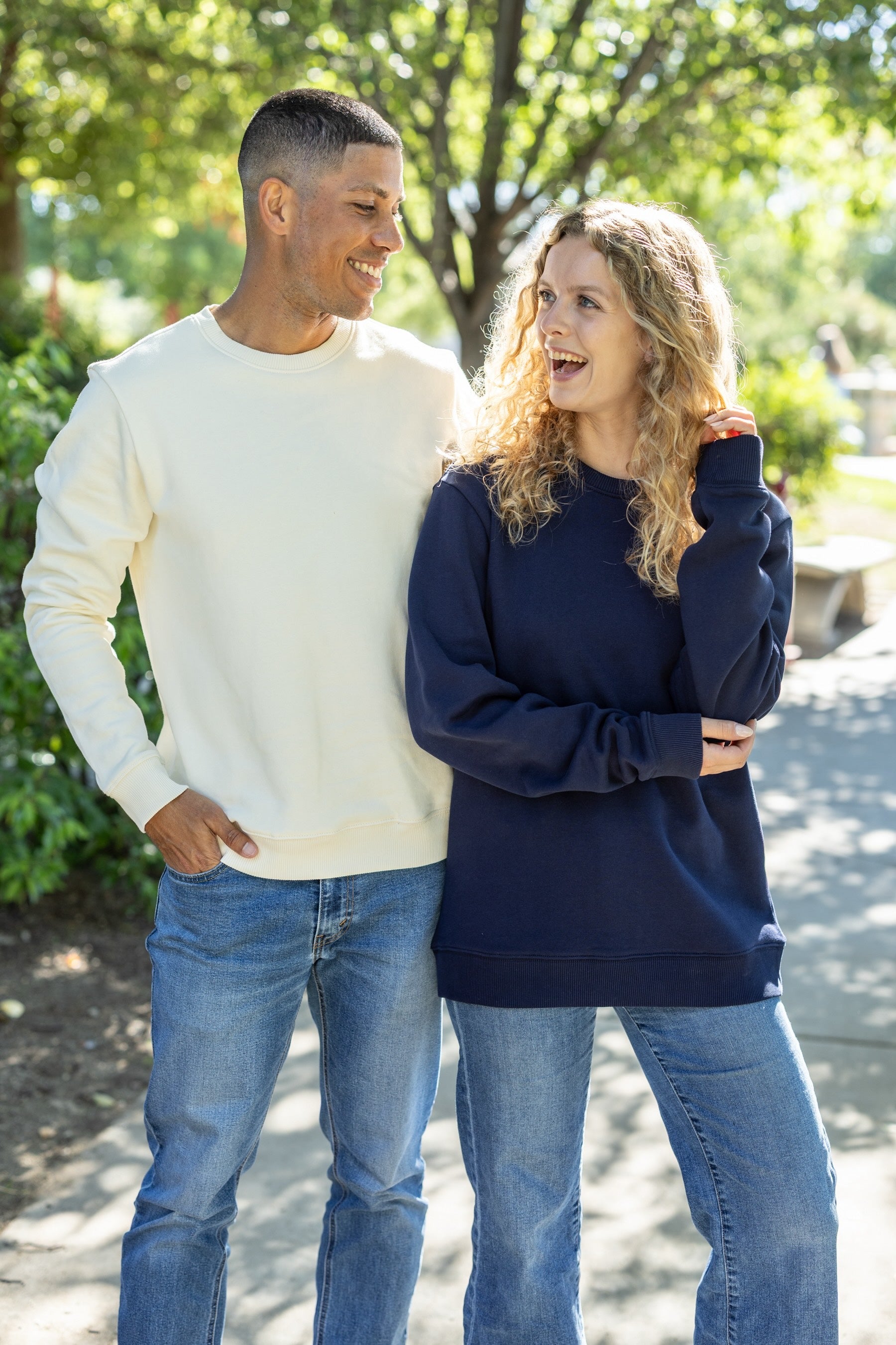 Two people standing outdoors, one wearing a white sweatshirt and jeans, the other in a navy sweatshirt and jeans.